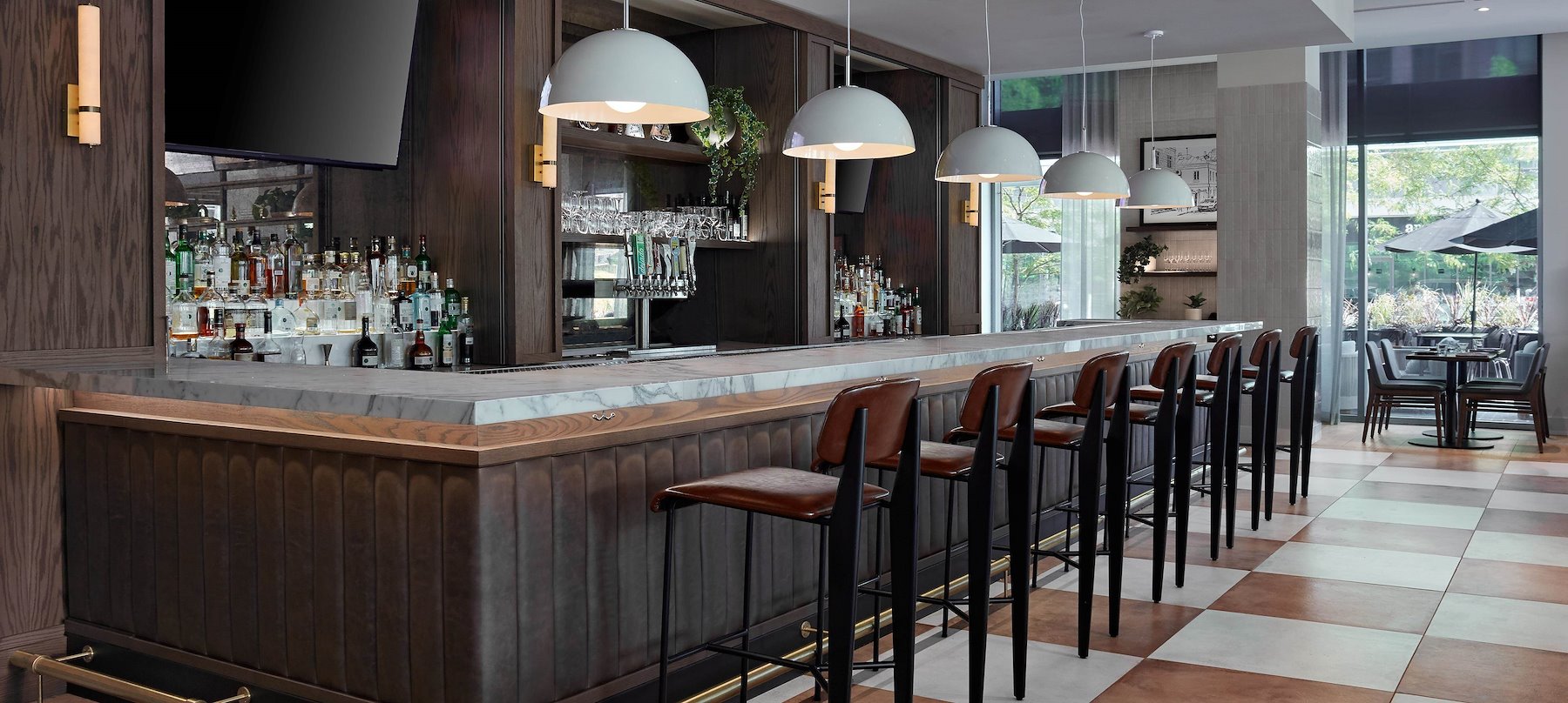 Modern bar with high chairs, pendant lights, TVs, and sunlit seating by large windows.